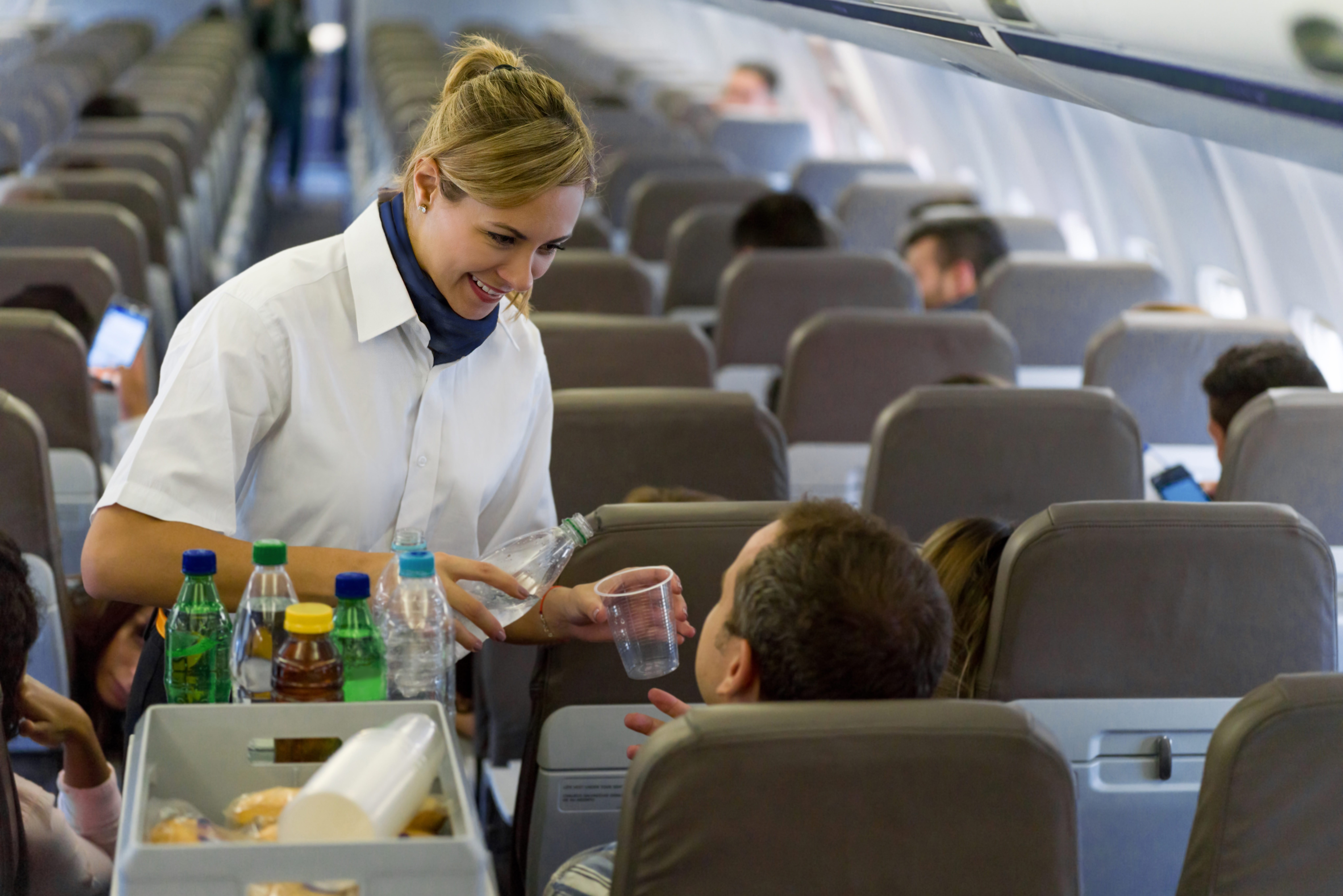 Flight attendant serves water to a passenger in an airplane aisle, with other passengers seated and occupied in a modern cabin environment.