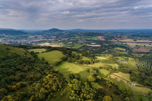 Rolling hills extend under a cloudy sky, revealing lush green fields, scattered trees, and a distant town nestled within the expansive landscape.