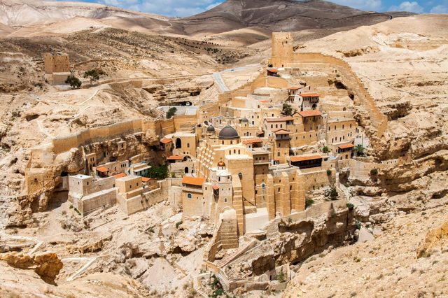 Stone buildings form a large, ancient monastery complex nestled into a rocky, arid desert landscape with hilltops and a blue sky.