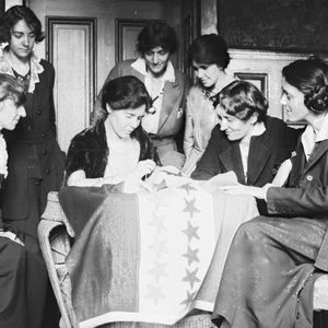 Seven women gather, sewing stars onto a banner, seated indoors with wooden paneling, sharing a collaborative atmosphere.