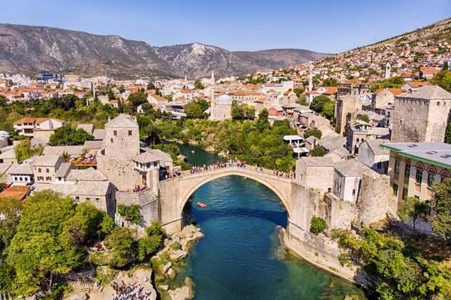 Stone bridge arches over a turquoise river, surrounded by historic buildings and greenery, set against a backdrop of mountains and cityscape under a clear sky.
