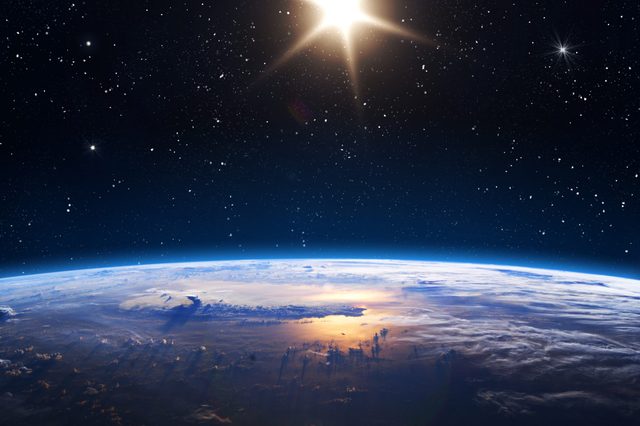 The Earth is partially illuminated, reflecting sunlight, against a backdrop of a starry sky and the sun shining brightly.