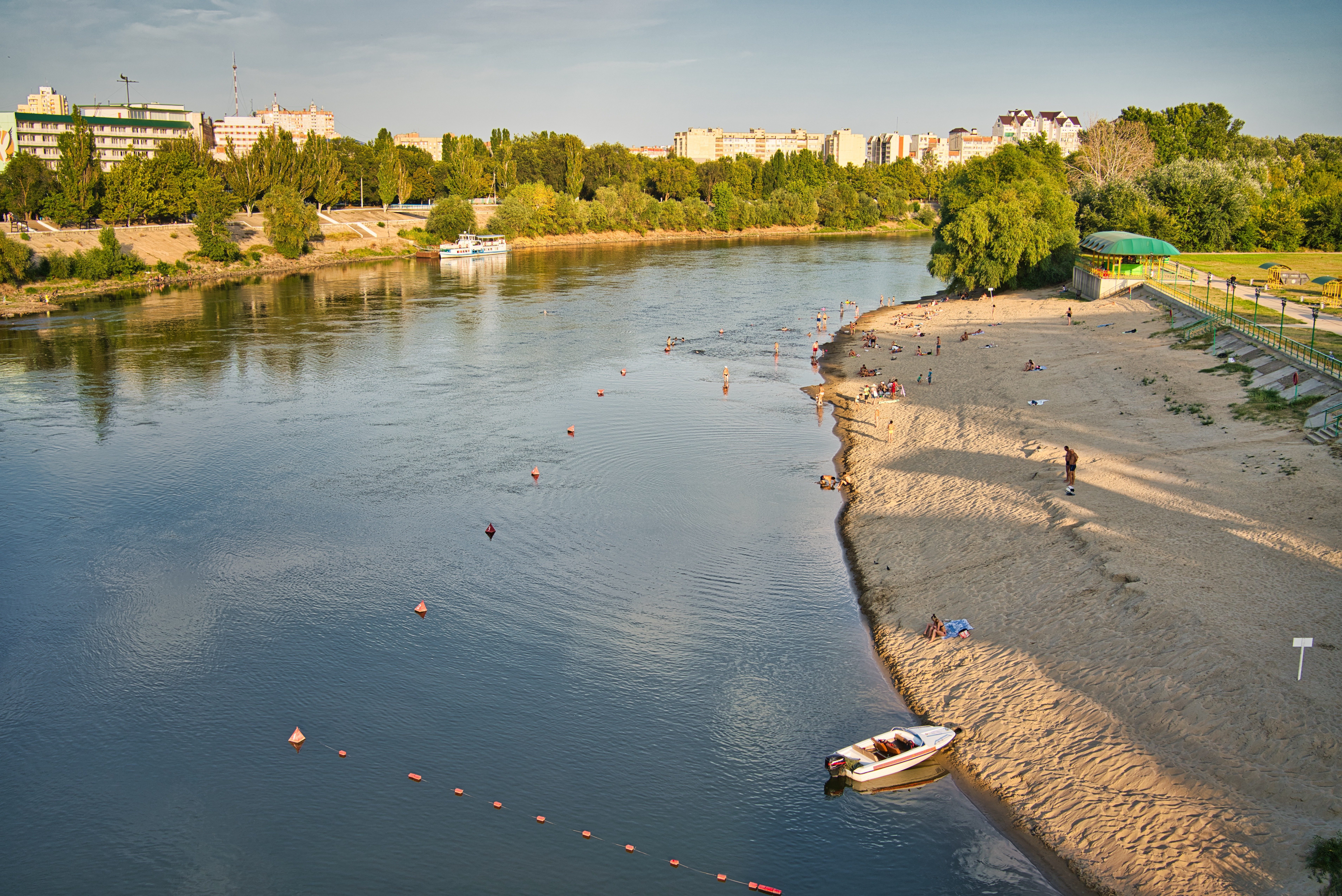 Beachgoers relax on a sandy riverbank, swimming near anchored boats, with green trees and city buildings lining the background.