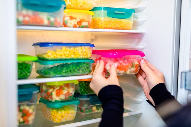Hands place colorful plastic containers filled with mixed vegetables into a refrigerator.