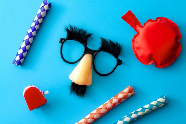 Novelty glasses with fake nose and mustache, surrounded by finger traps, wind-up chattering teeth, and a whoopee cushion on a blue surface.