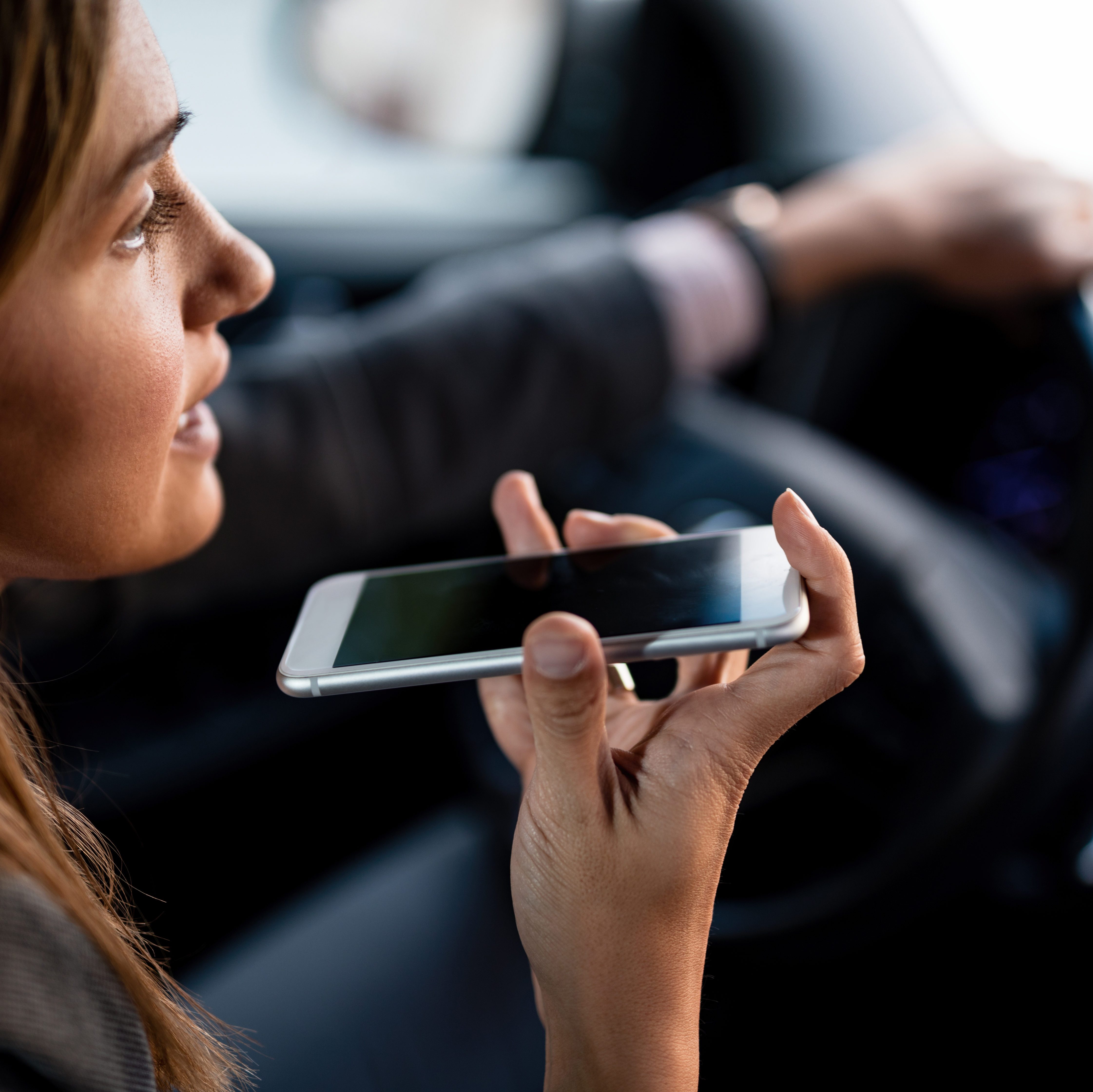A person holds a smartphone, speaking into it, while driving a car, with one hand on the steering wheel.