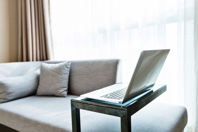 Laptop sits open on a glass table beside a gray sofa, with soft lighting from sheer curtains in a modern living room.