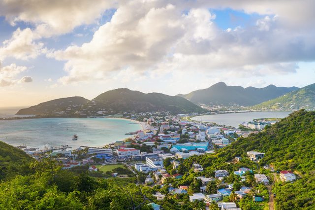 A coastal town sits between lush hills, overlooking a bright bay under a partly cloudy sky and soft sunlight.