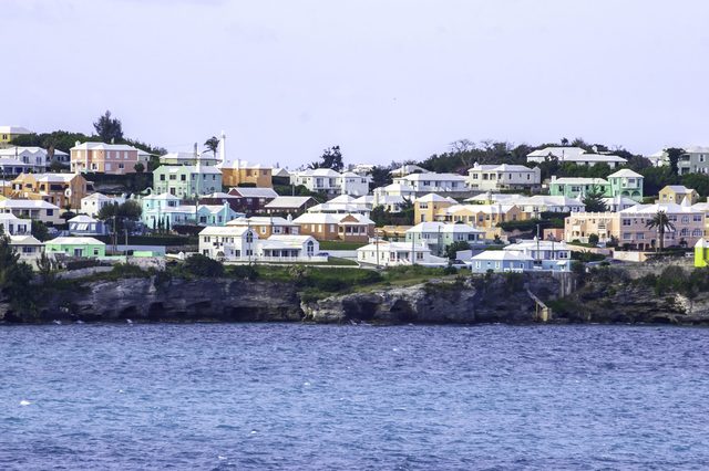 Colorful houses stand on a rocky coastline, surrounded by lush greenery and overlooking a calm ocean under a clear sky.