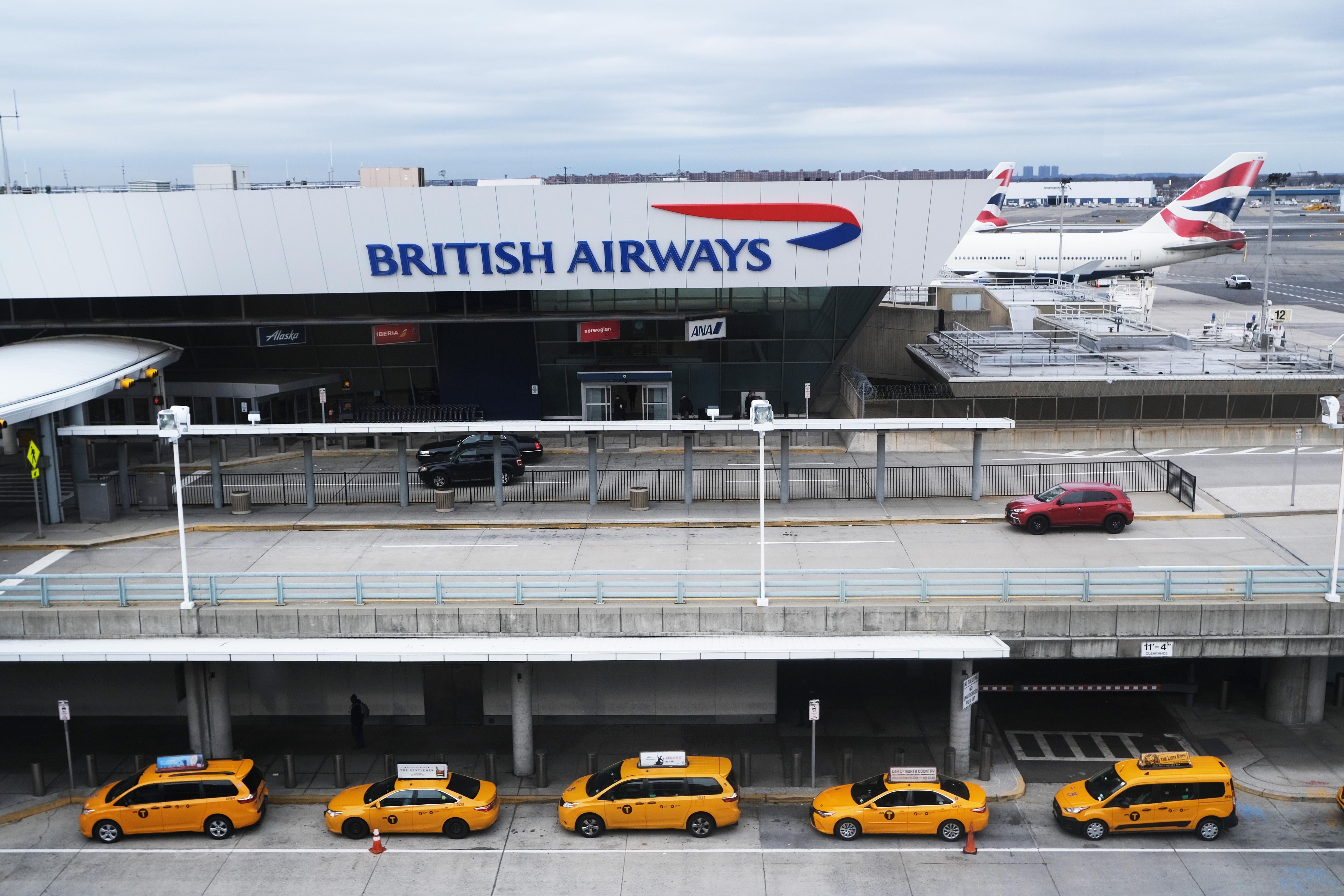 Taxis stand parked in a row outside a British Airways terminal at an airport, with a plane visible on the tarmac behind.