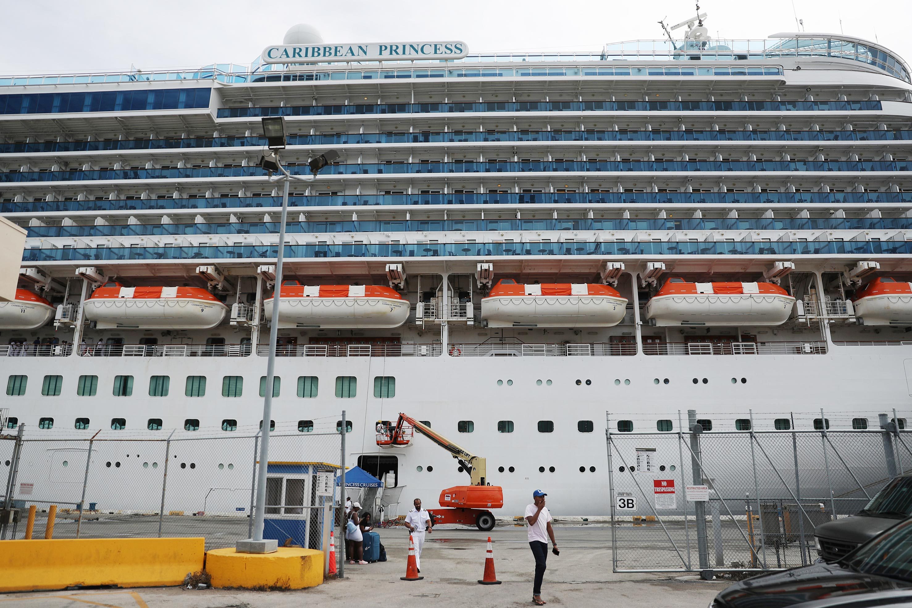 Cruise ship docked, people conversing at port; lifeboats visible; fenced area with parked cars. Sign reads "CARIBBEAN PRINCESS" on the ship.