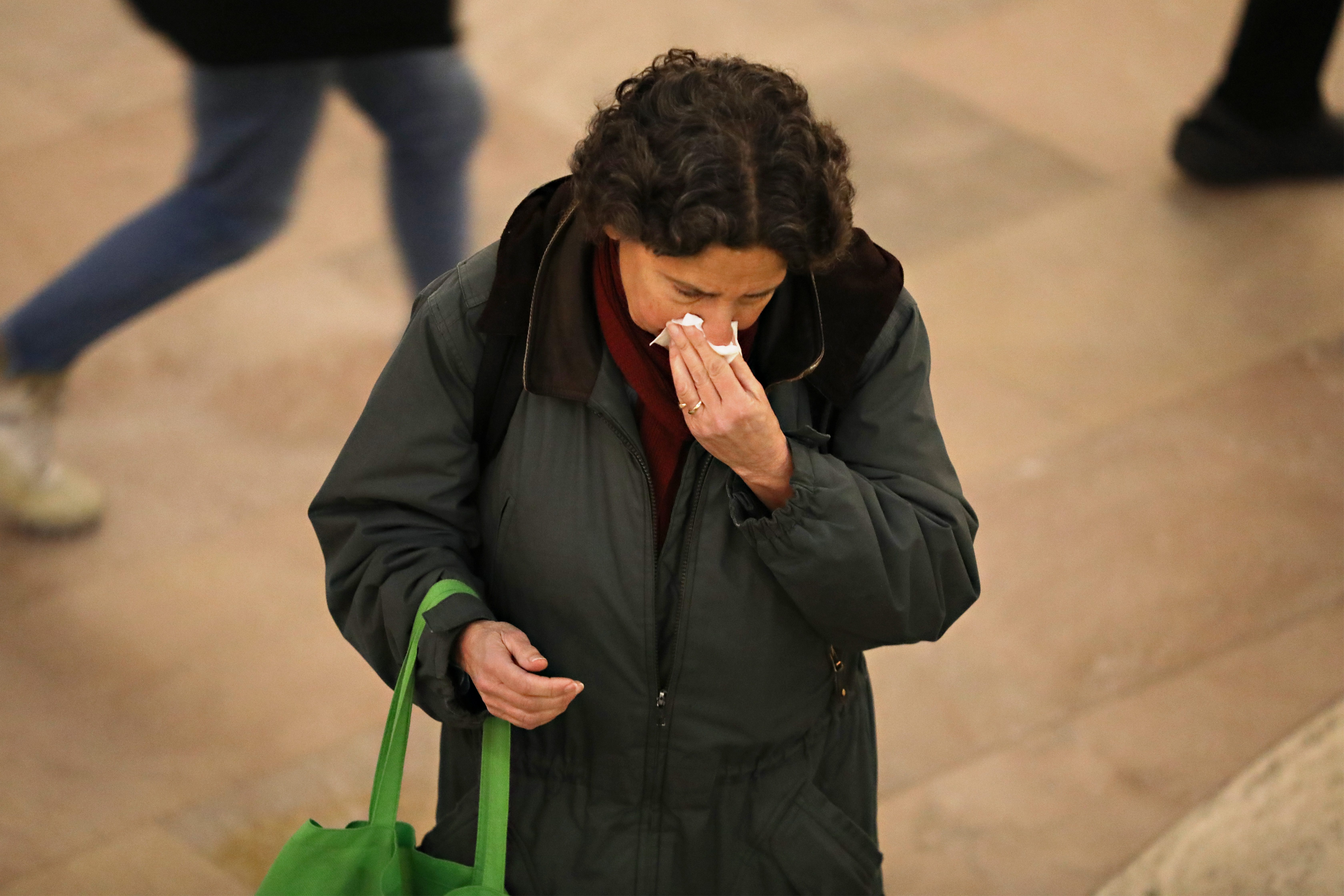 Person holding tissue to nose, walking indoors, wearing a green jacket, and carrying a bright green bag. Other people walk past on a tiled floor.