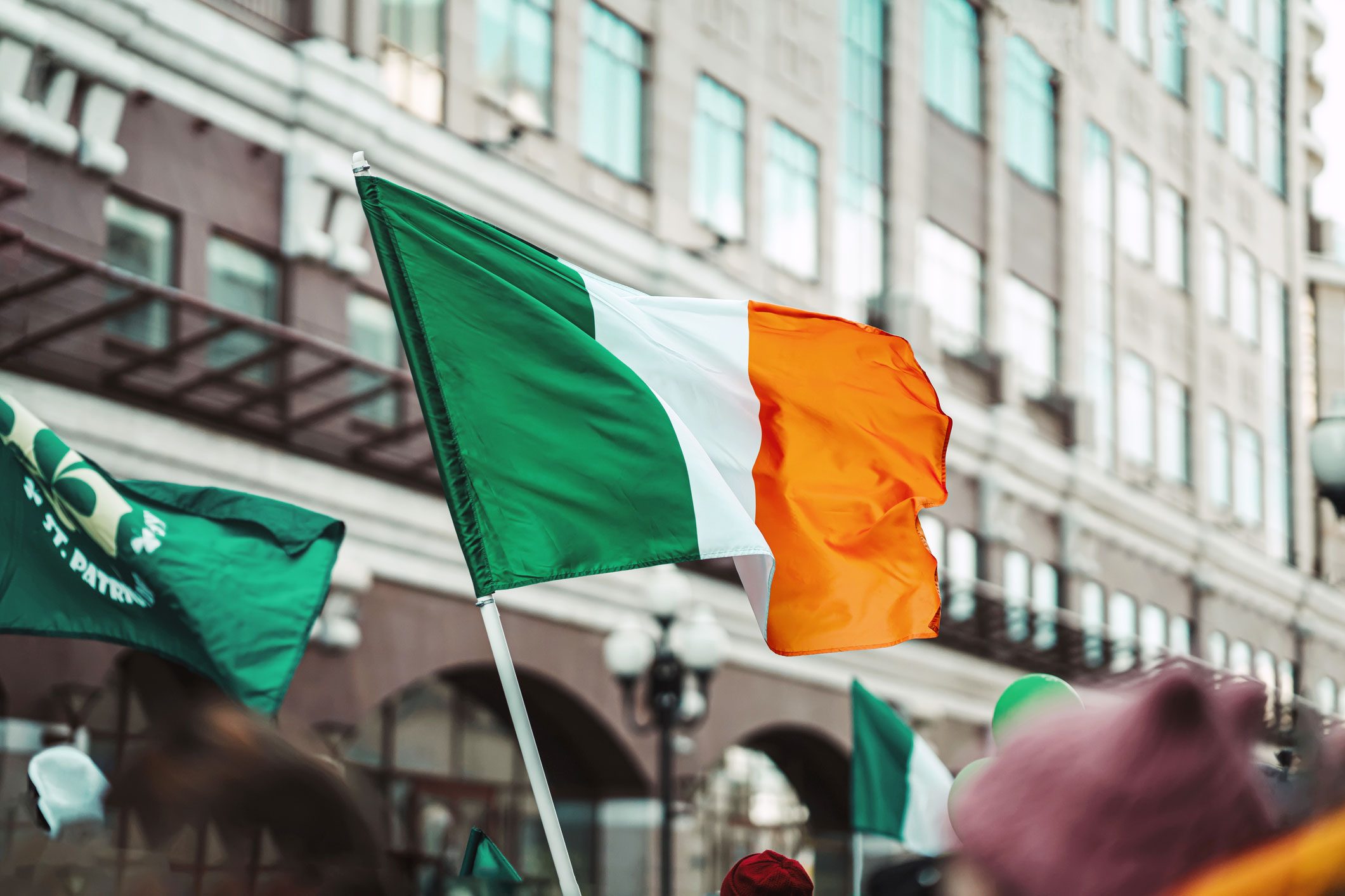 Irish flag waves amidst a city parade with blurred participants and buildings in the background.