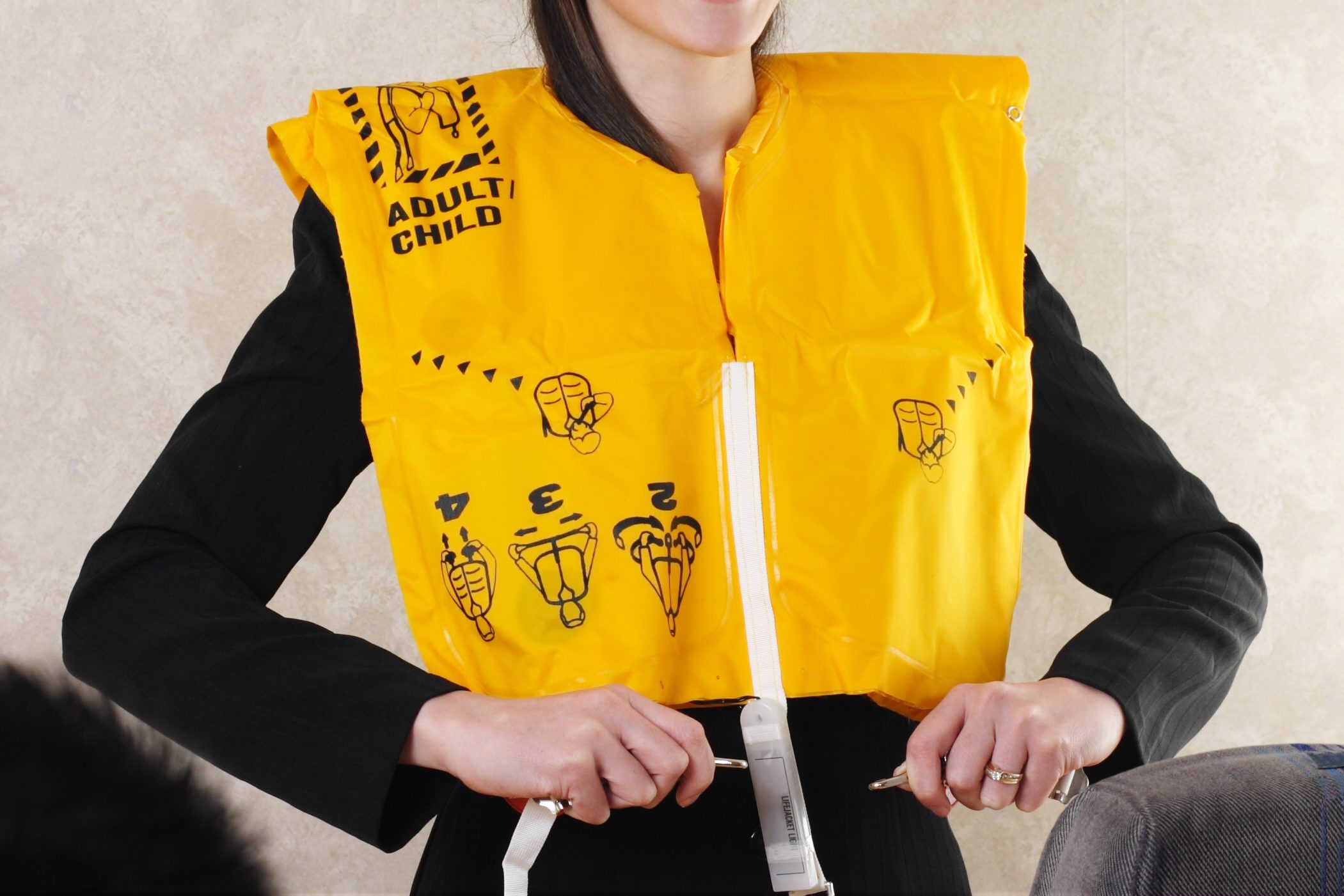 A person demonstrates wearing a yellow life jacket labeled "ADULT/CHILD" with illustrated instructions, while standing indoors near a seat.