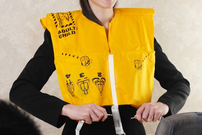 A person demonstrates wearing a yellow life jacket labeled "ADULT/CHILD" with illustrated instructions, while standing indoors near a seat.