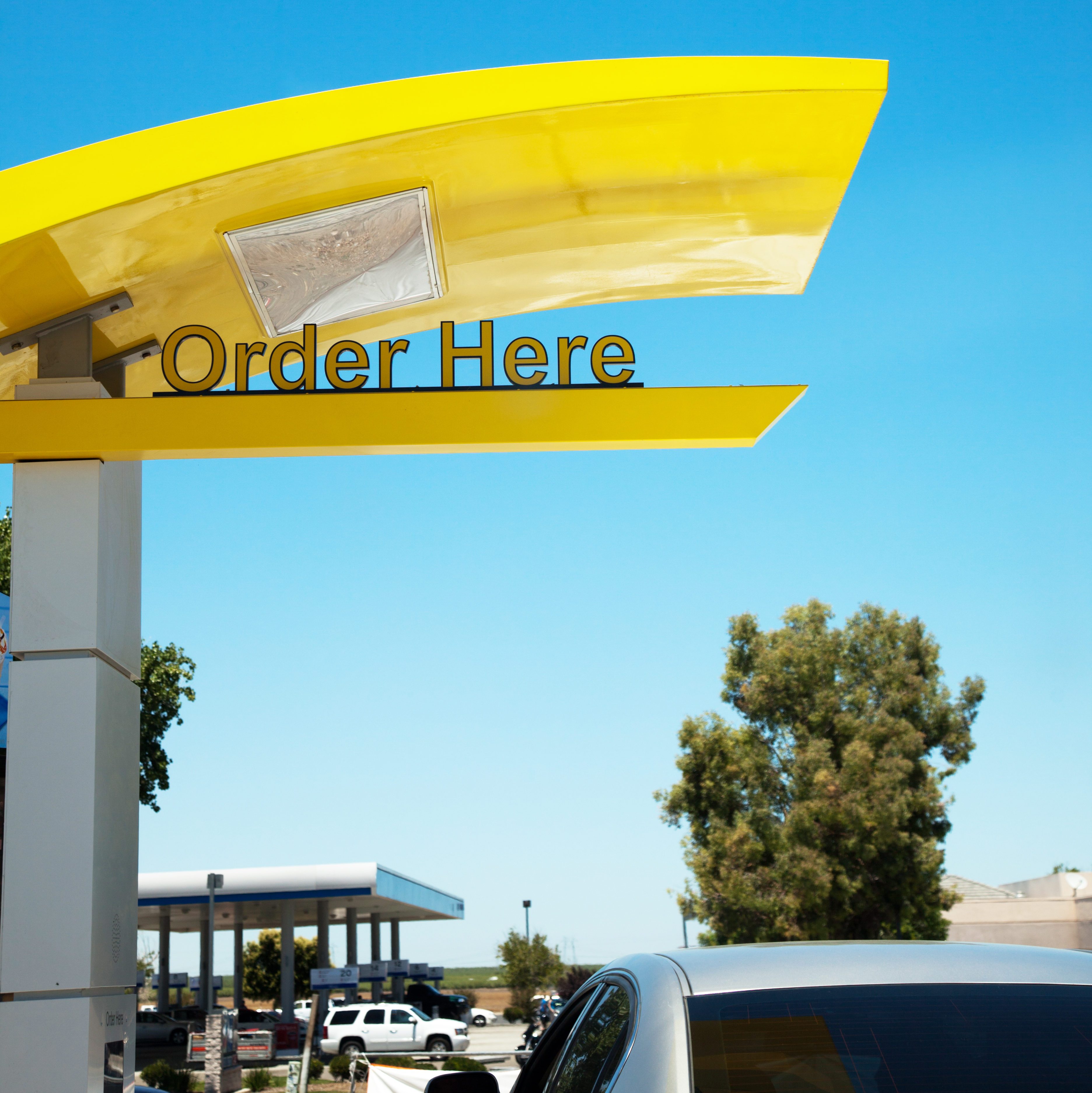 Drive-thru order station displays "Order Here" under yellow canopy, with cars and trees visible in the sunny outdoor setting.