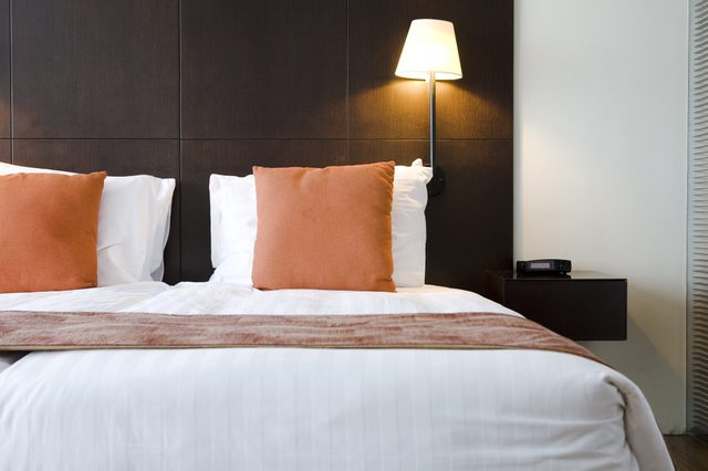 Bed with white linens and two orange pillows under a lit wall lamp; dark headboard, bedside table with a clock in a modern bedroom.