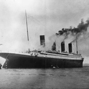Large ship emits smoke, moving across water alongside smaller tugboats, under an overcast sky.