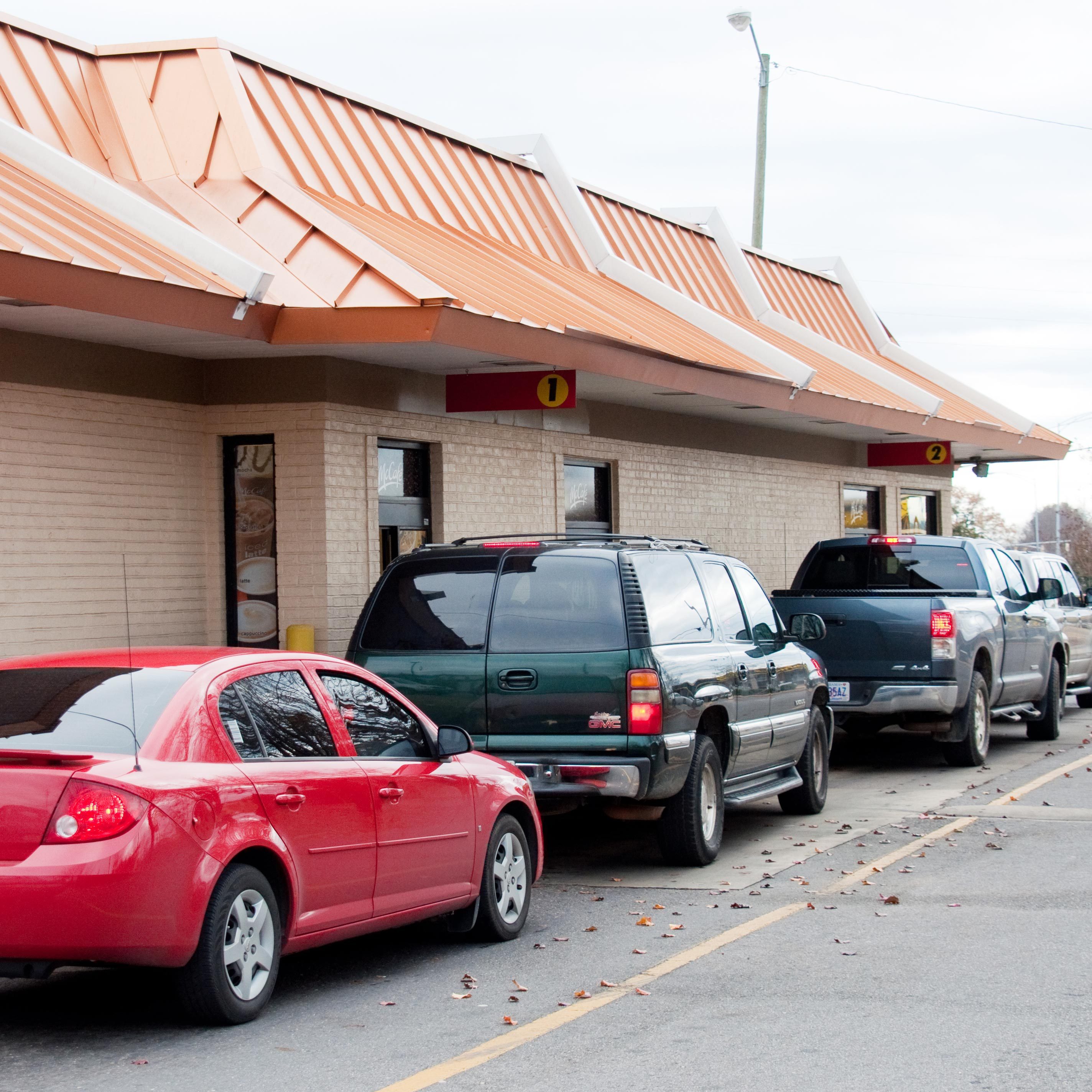 Cars lined up at a drive-thru under an orange roof, waiting for service. The background shows a clear sky and scattered leaves.