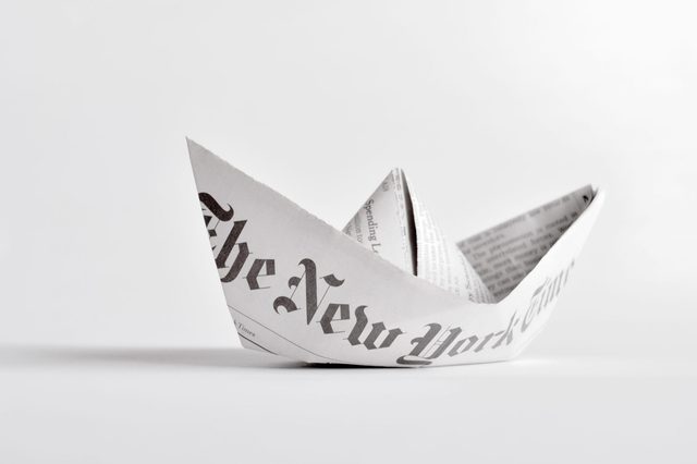 Paper boat sits on flat surface, folded from newspaper with "The New York Times" text partially visible, against a plain white background.