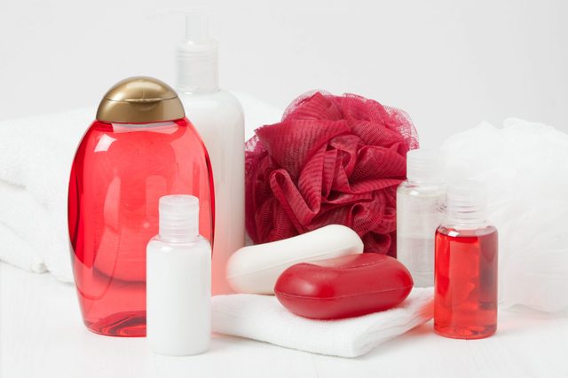 Bath products rest on towels; includes red and white bottles, soaps, and a red shower pouf in a bathroom setting.