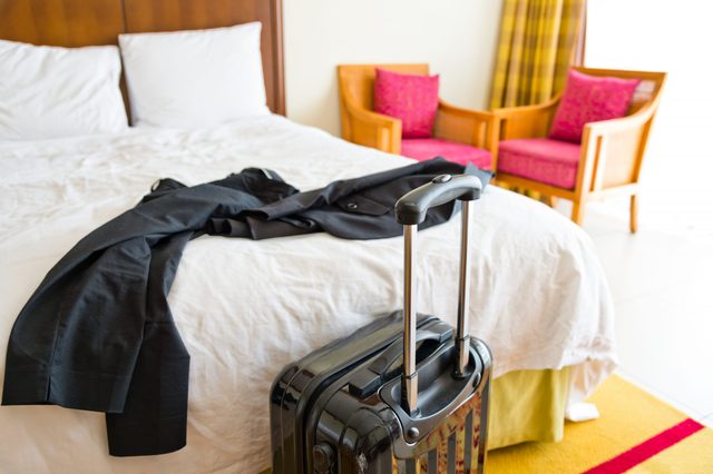 A suitcase stands next to a bed with a neatly laid suit jacket in a bright hotel room with chairs and curtains.