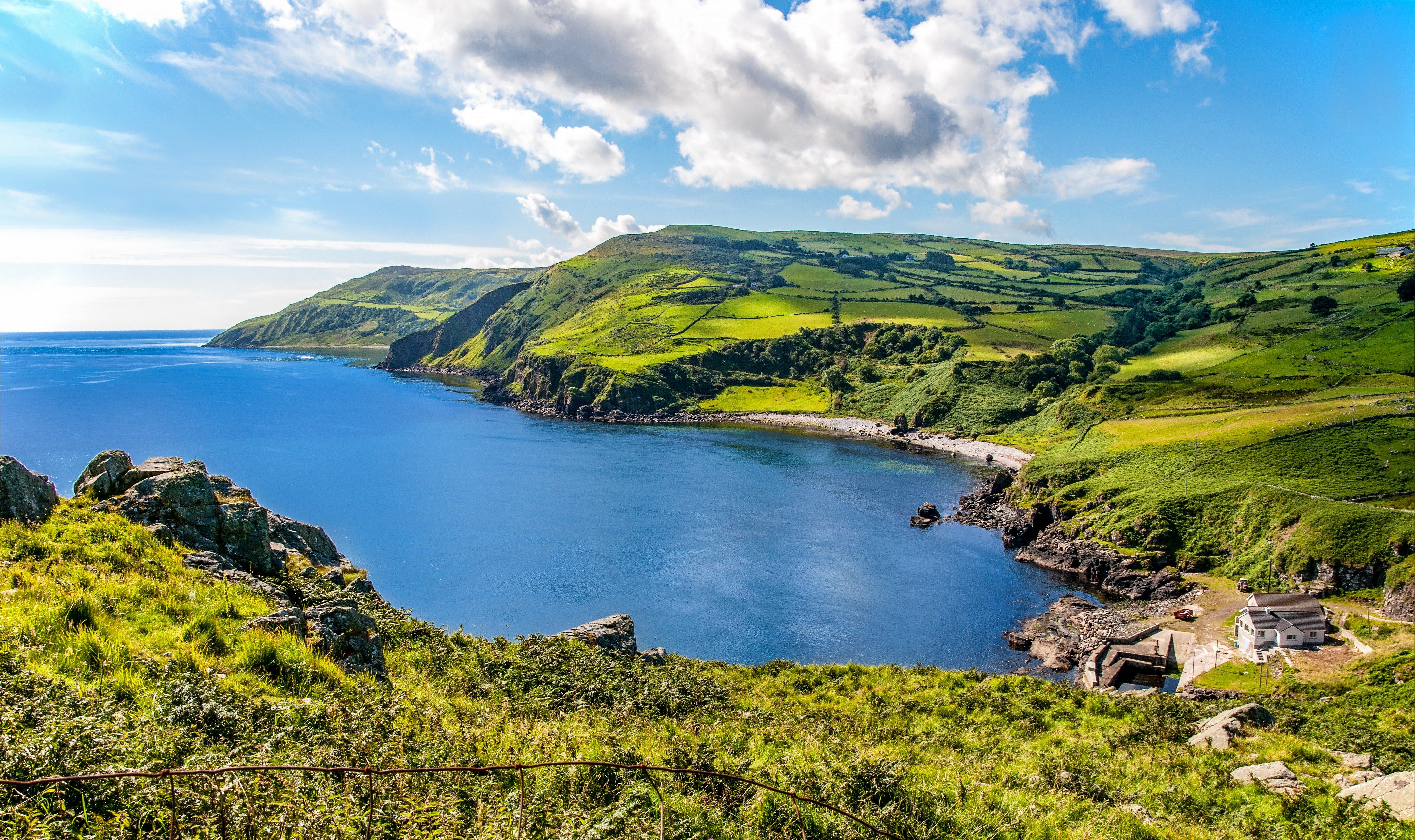 Coastal landscape displays lush green hills meeting a calm blue sea under a bright sky with scattered clouds. A small house sits near the shore.