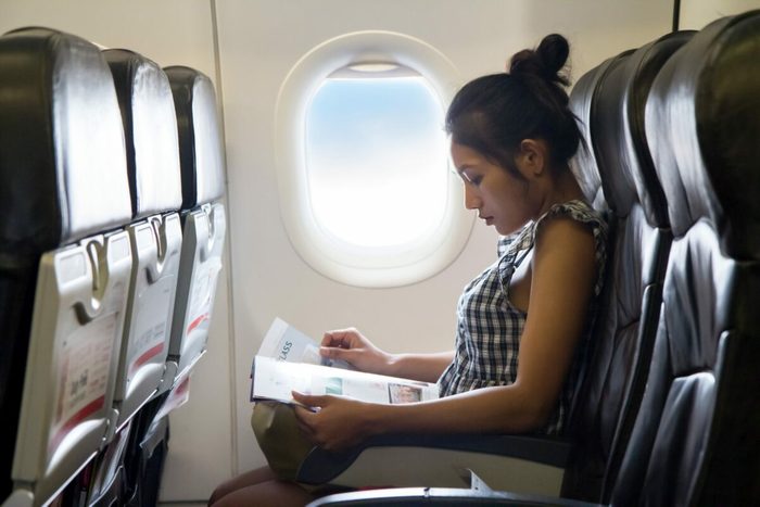 Person reading a magazine while sitting on an airplane, sunlight streaming through the window, surrounded by empty seats.