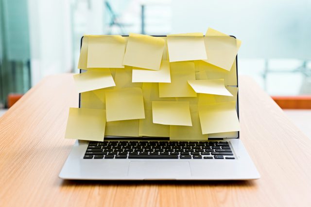 Laptop covered with yellow sticky notes sits unused on a wooden desk in a modern, brightly lit office environment.