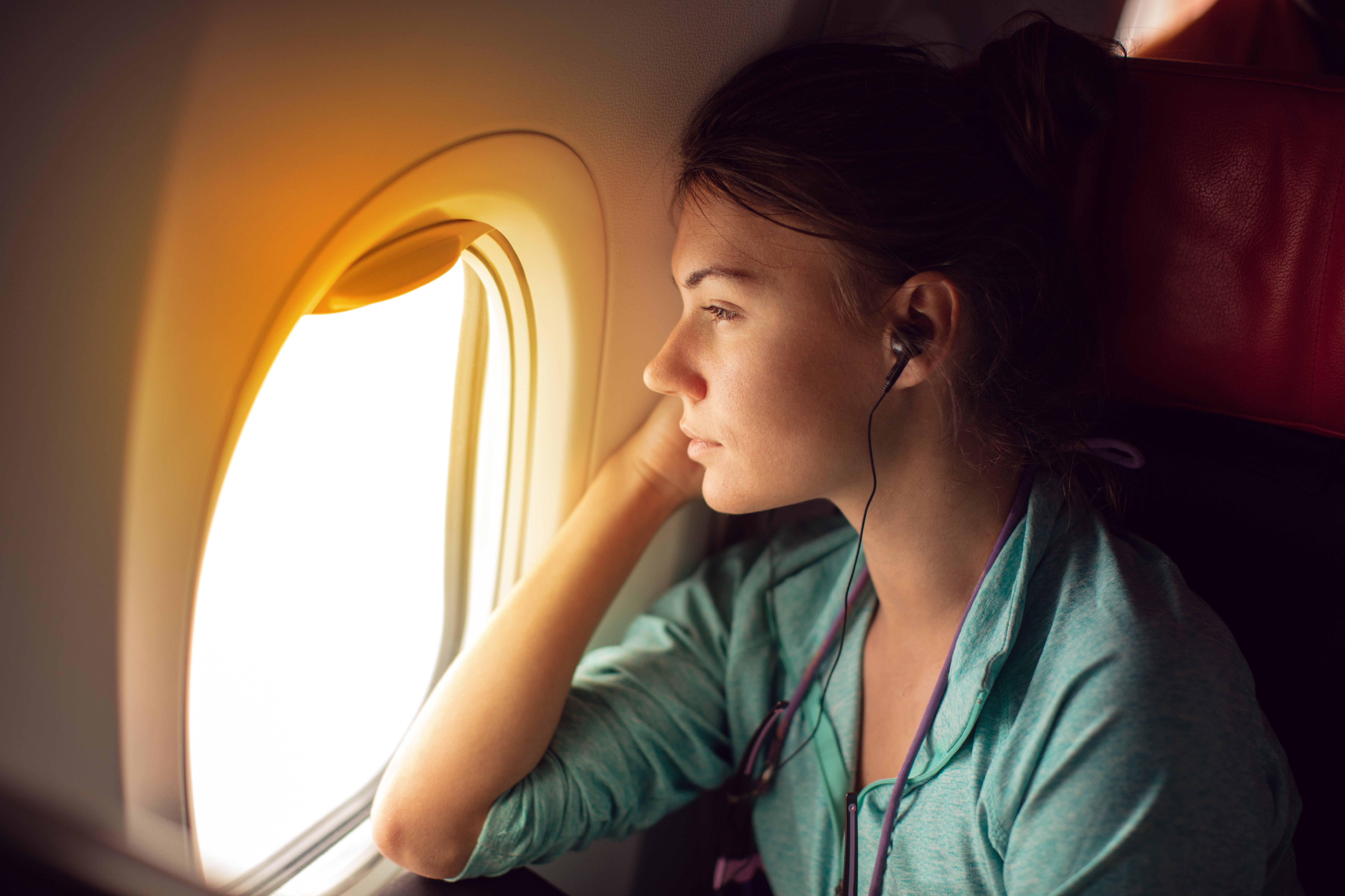 Person with headphones gazing out airplane window, sitting in a seat, illuminated by soft, warm light.