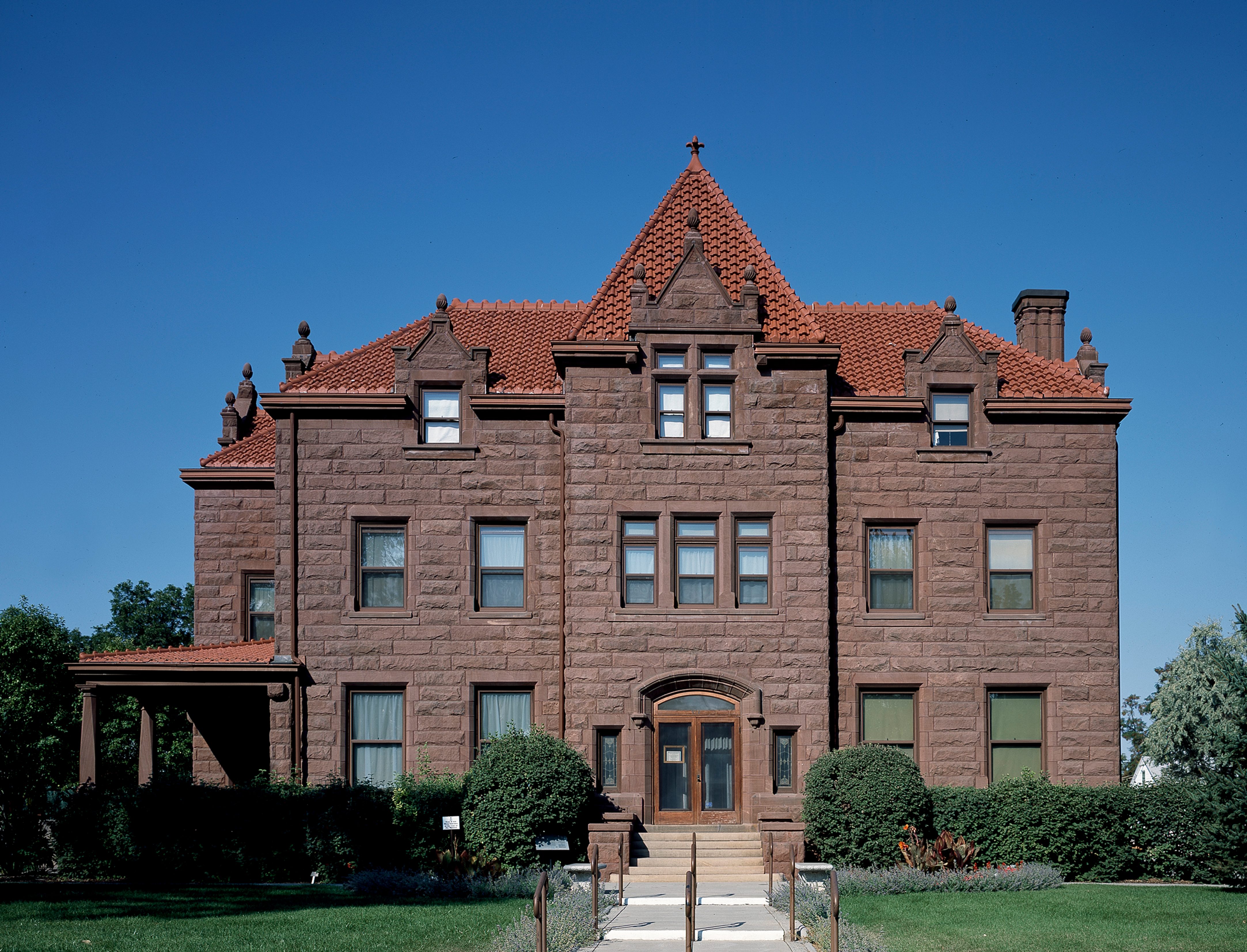 A large, stone mansion stands with a terracotta-tiled roof under a clear blue sky, surrounded by green shrubs and a neatly kept lawn.