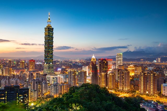 Skyscraper towers over a vibrant cityscape at twilight, surrounded by illuminated buildings and distant mountains beneath a clear, blue sky.