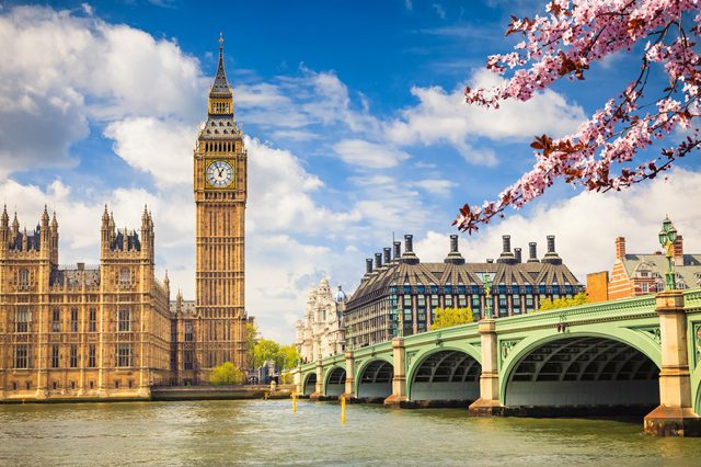Clock tower stands tall beside river; cherry blossoms frame scene; adjacent buildings and bridge under bright sky.