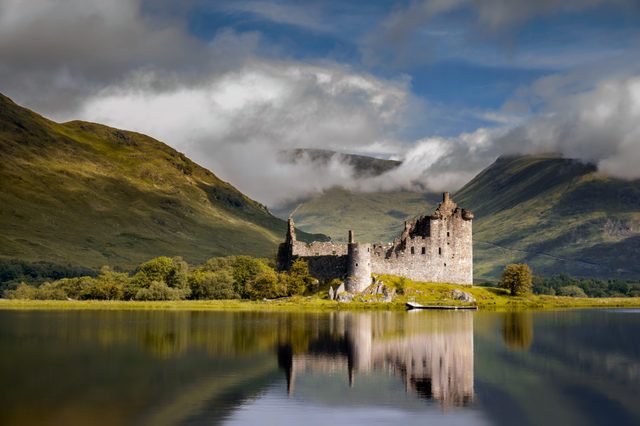 Castle ruins reflect on a calm lake, surrounded by lush green hills under a cloudy sky.