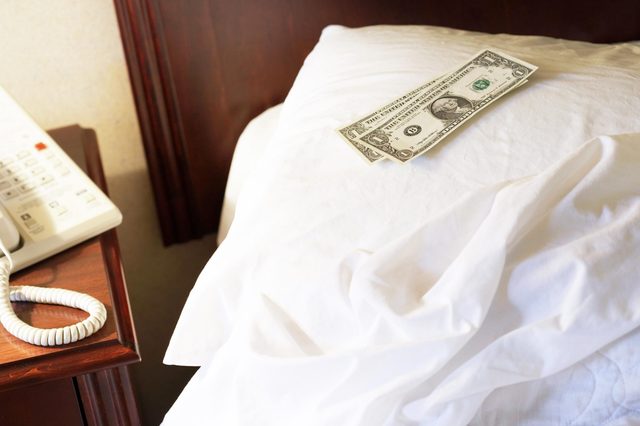 Dollar bills resting on a pillow, beside a wooden bedside table with a telephone, indicating a hotel room setting.