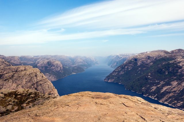 Rocky cliff overlooks a wide, calm fjord surrounded by steep mountains under a clear blue sky.