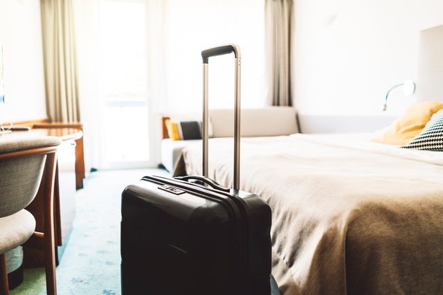 Suitcase stands upright beside a bed in a sunlit hotel room with modern furnishings and a window.