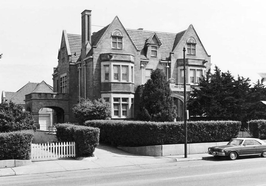 A large, historic brick house sits behind hedges and a white picket fence, with a parked car beside it on a suburban street.