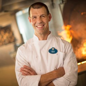Chef stands smiling, arms crossed, with flames in the background. Name tag reads 