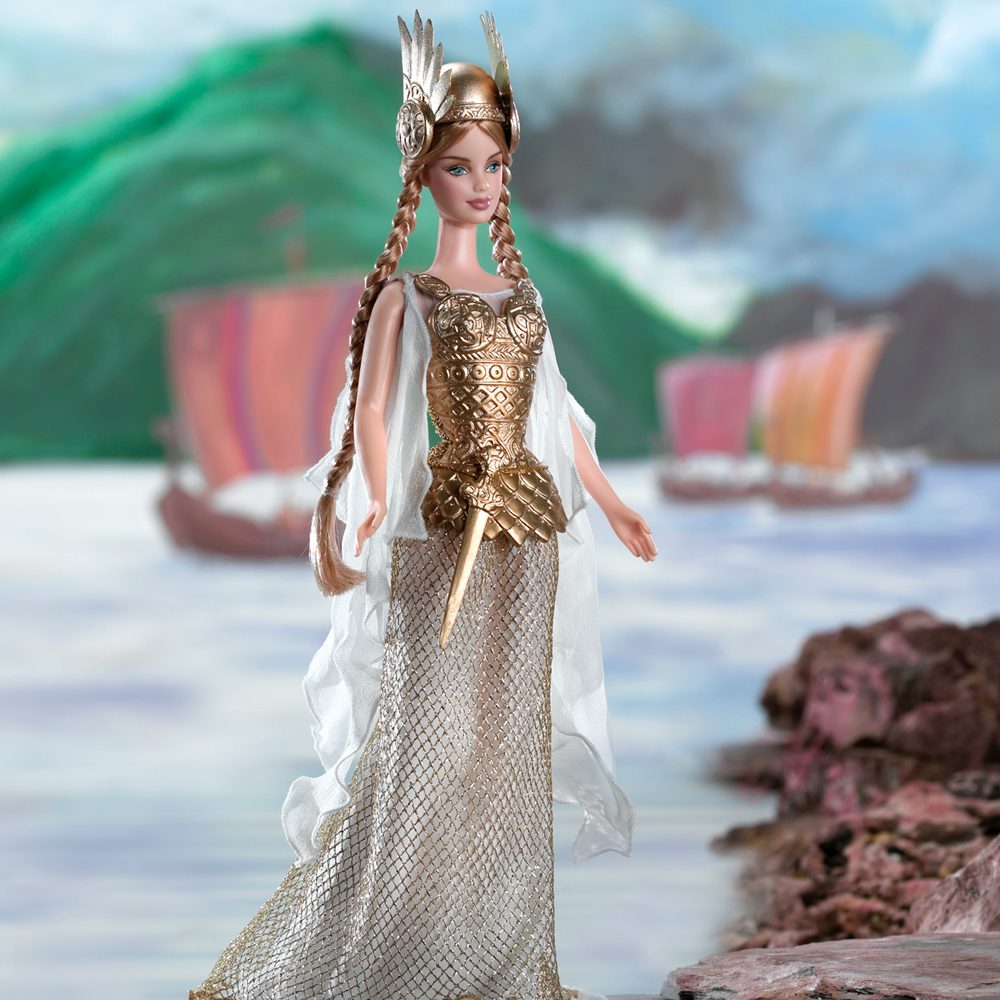 Doll wearing golden armor and winged helmet stands confidently near a rocky shore, with colorful sailing ships in the background.
