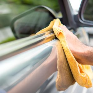 Hand wipes car with yellow cloth, reflecting in shiny surface outdoors.