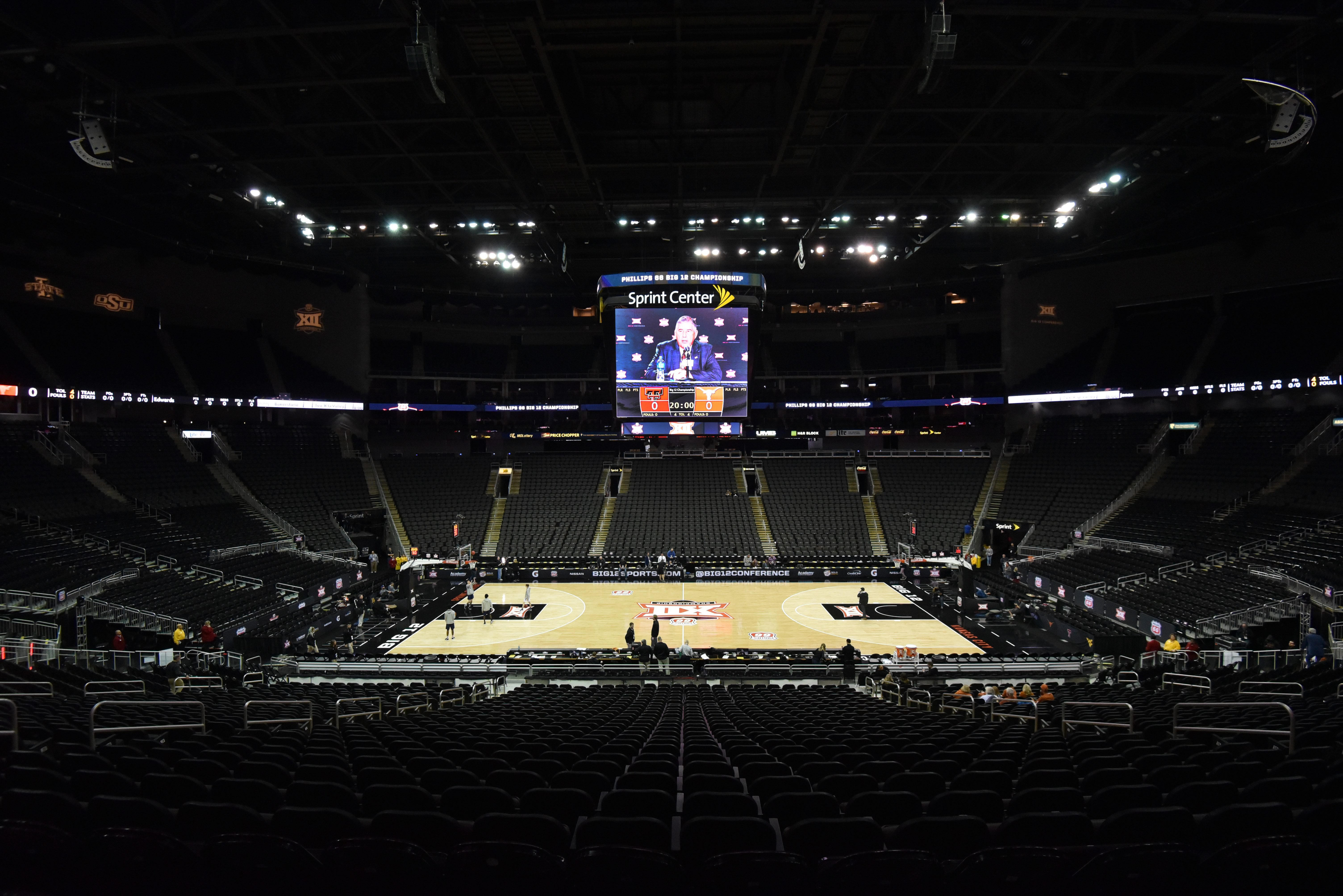 Basketball court lies empty, surrounded by rows of seats in a dimly lit arena. A large screen displays a speaker above the court.