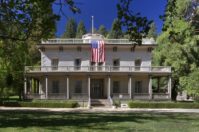 A grand, two-story stone house displays an American flag on its balcony, surrounded by trees and a grassy lawn.
