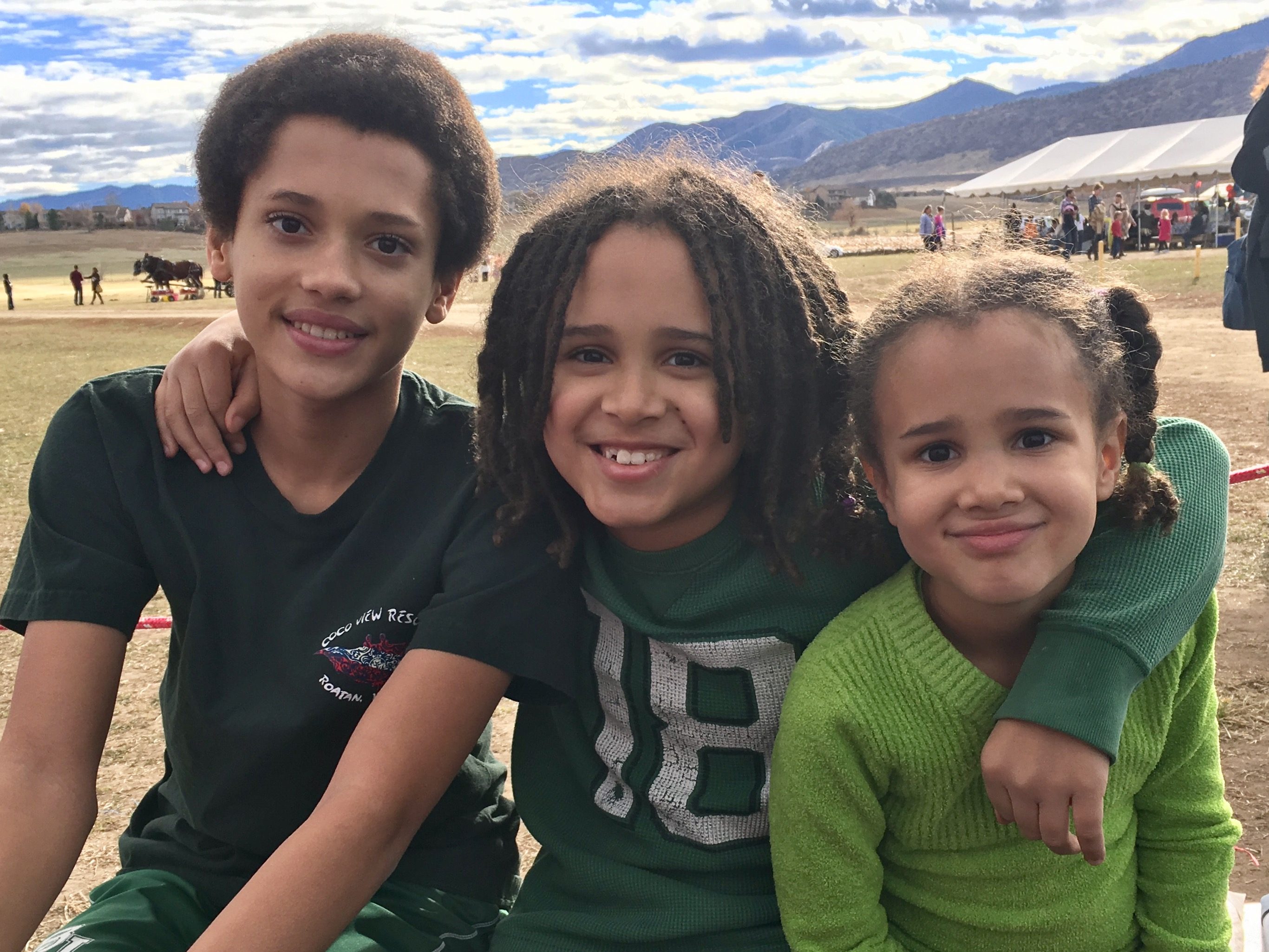 Three children smile seated outdoors; behind them are fields, distant mountains, and a tent with people.