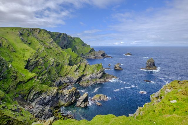 Cliffs covered in green grass overlook a blue ocean with scattered rocks, beneath a partly cloudy sky.