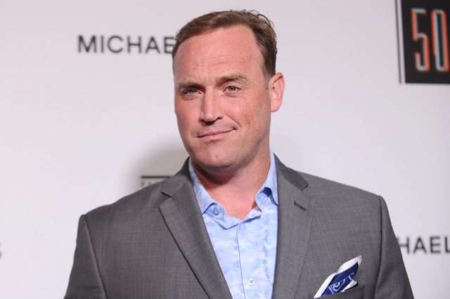A person stands, wearing a gray suit and a patterned shirt, against a backdrop with partial text "MICHAEL" and "50".