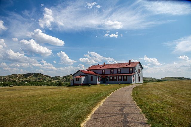 A large house with red roof stands surrounded by green fields and rolling hills under a partly cloudy blue sky.