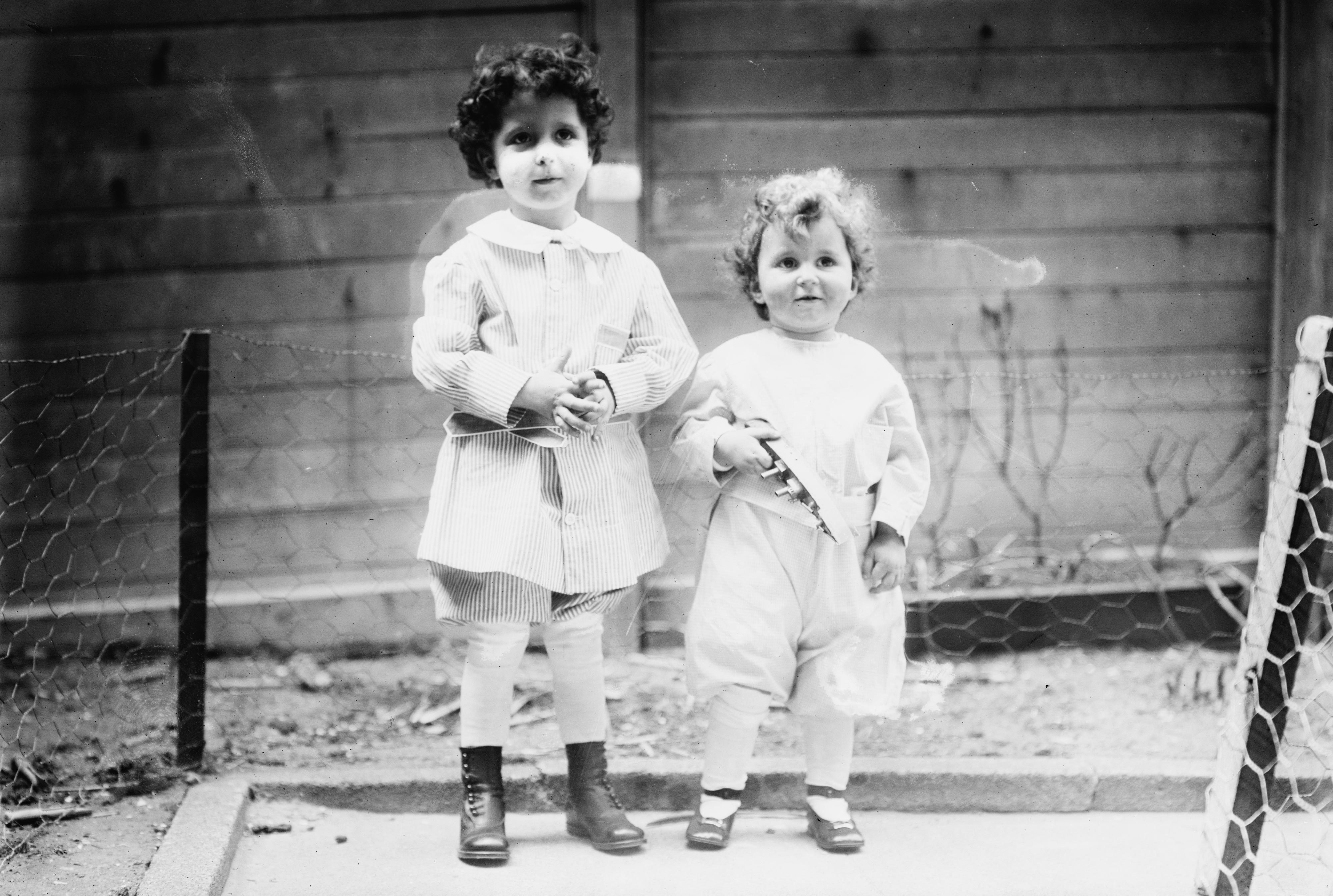Two children stand together, one holding a toy, in a garden with a wooden fence and wire mesh surrounding them.