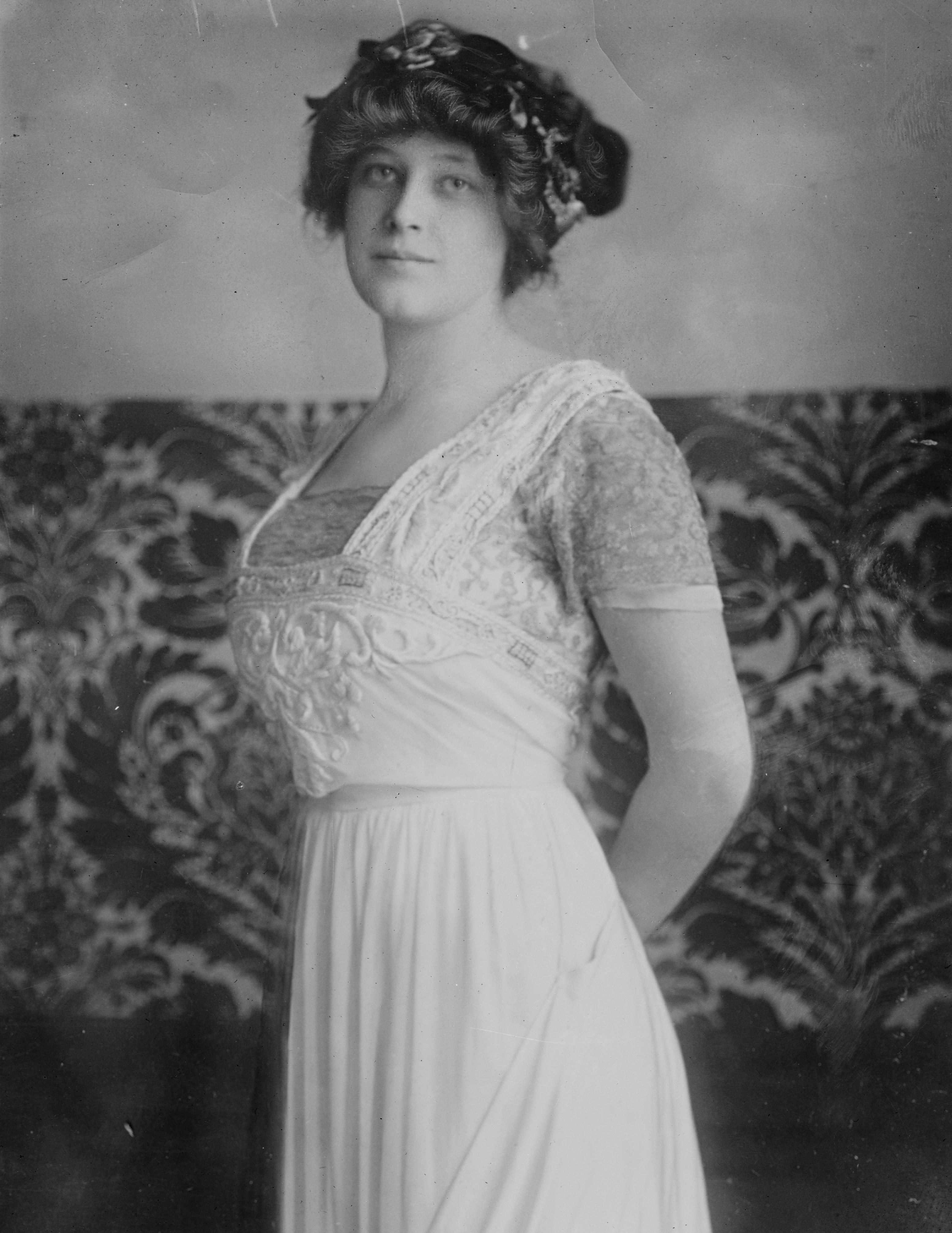Woman stands, wearing an embroidered gown and floral headpiece, against a patterned backdrop, evoking early 20th-century elegance.