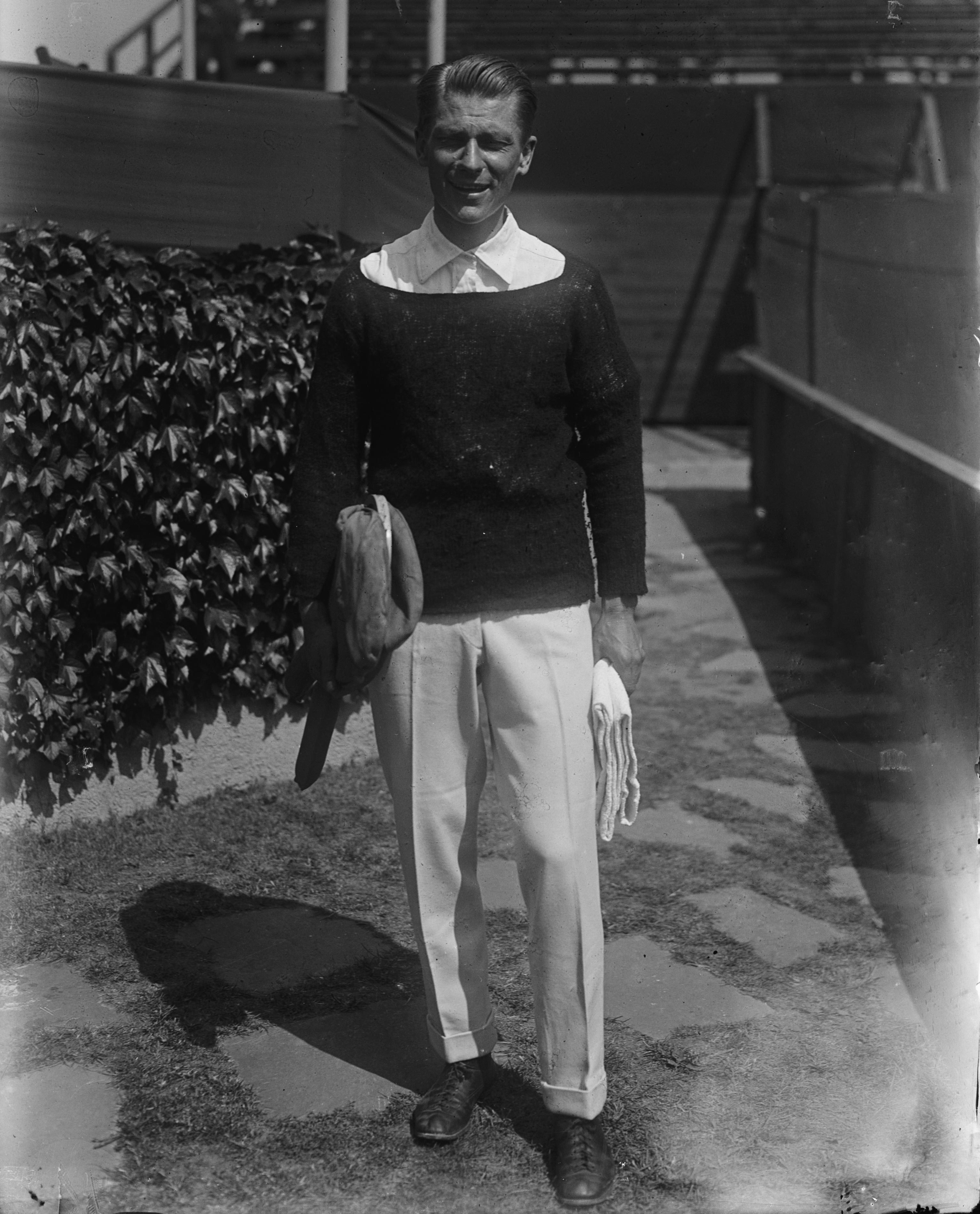 A person stands holding a towel and racket, dressed in a sweater and white pants, near a vine-covered wall in an outdoor setting.